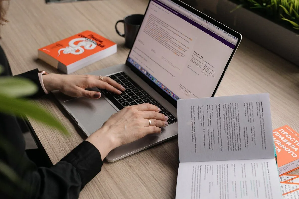 Person browsing the web on a laptop while reading a book at a desk